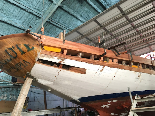 A classic wooden sailing boat inside a workshop, undergoing maintenance and restoration.